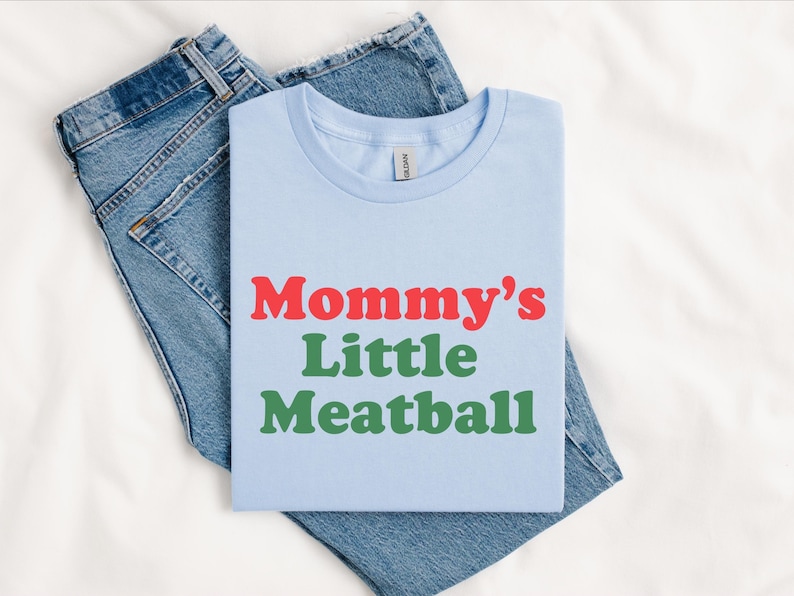 May include: Pale blue t-shirt featuring the text "Mommy's Little Meatball" in red and green. The t-shirt is folded and placed on a pair of blue denim jeans.