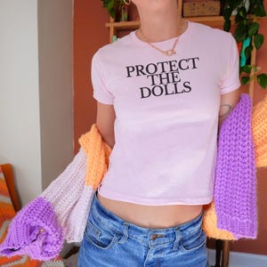 Puede incluir: Una camiseta rosa claro con el texto negro "PROTECT THE DOLLS". La persona lleva vaqueros azules y sostiene un c&aacute;rdigan de punto colorido con secciones naranjas, moradas y blancas. El fondo incluye una silla y una estanter&iacute;a.