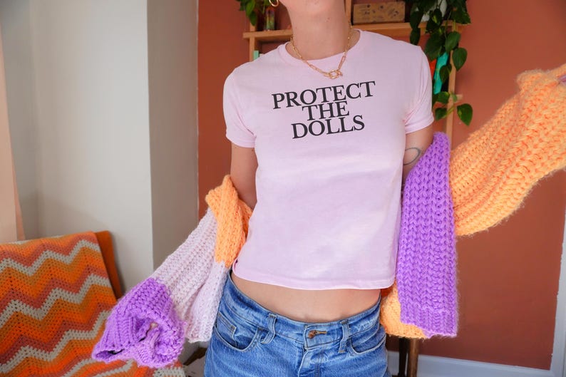 Puede incluir: Una camiseta rosa claro con el texto negro "PROTECT THE DOLLS". La persona lleva vaqueros azules y sostiene un c&aacute;rdigan de punto colorido con secciones naranjas, moradas y blancas. El fondo incluye una silla y una estanter&iacute;a.