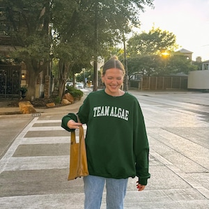 May include: A dark green crewneck sweatshirt featuring the white text TEAM ALGAE printed in an arched font across the chest, paired with light blue denim jeans and a tan leather tote bag held at the side, creating a casual everyday outfit.