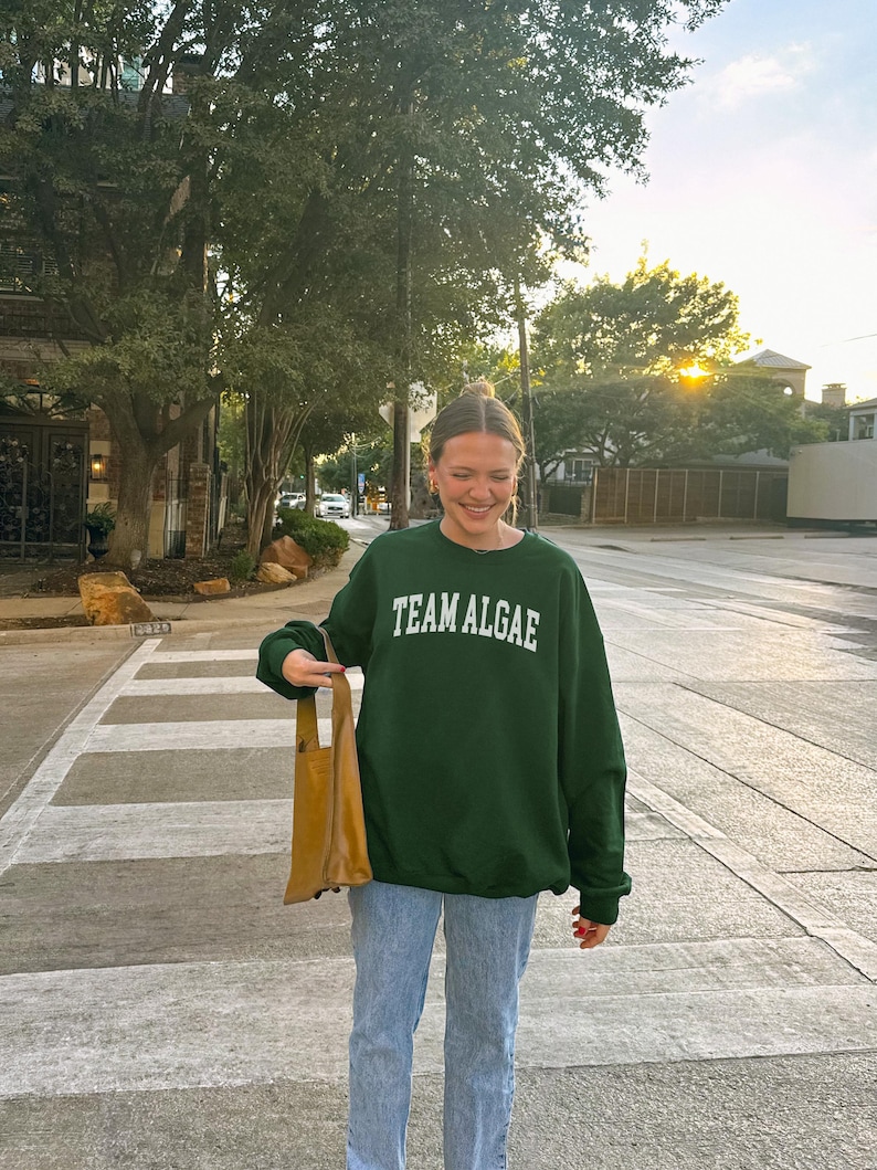 May include: A dark green crewneck sweatshirt featuring the white text TEAM ALGAE printed in an arched font across the chest, paired with light blue denim jeans and a tan leather tote bag held at the side, creating a casual everyday outfit.