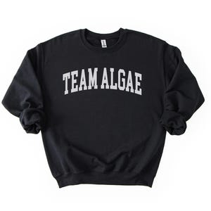 May include: A black crewneck sweatshirt featuring the text "TEAM ALGAE" printed in a white, arched, collegiate-style font across the chest. The garment has long sleeves with ribbed cuffs and a ribbed hem, providing a standard fit and silhouette.