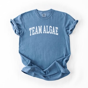May include: A blue short-sleeve crew neck t-shirt with the text "TEAM ALGAE" printed in white capital letters across the chest in an arched font. The garment features a standard fit with cuffed sleeves and a ribbed collar neckline.