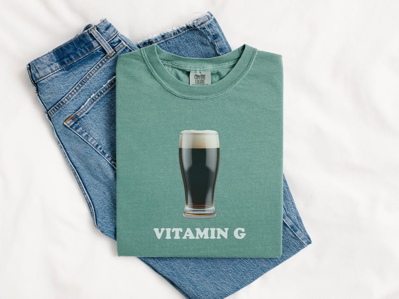 Pu&ograve; includere: Una maglietta verde con un bicchiere di birra scura e la scritta "VITAMIN G" stampata su di essa.