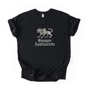 May include: Black t-shirt featuring a two-headed dog illustration in gray. The text "Siempre Antifascista" is printed below the illustration in a gothic font. The shirt is made of a soft material.