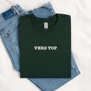 May include: A dark green t-shirt with the text "VERS TOP" printed in white on the front. The t-shirt is folded and placed on top of a pair of blue jeans.