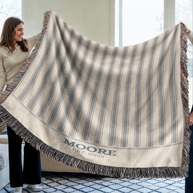 May include: A beige and gray striped woven throw blanket with a fringed edge. The blanket has vertical gray stripes and the word "MOORE" and the year "EST. 2025" are printed on the bottom edge.