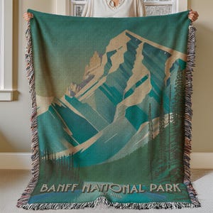 May include: A woven tapestry throw blanket featuring a vintage-style illustration of a mountain range with the text "Banff National Park Est. 1885". The blanket has a green, blue, and brown color scheme.