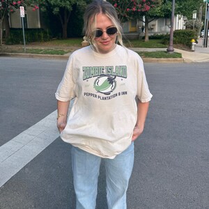 May include: A woman wearing a light beige t-shirt with a green graphic print that says "Zombie Island" and "Pepper Plantation & Inn". She is also wearing light blue jeans.