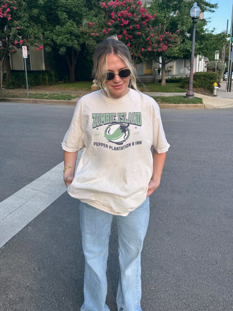May include: A woman wearing a light beige t-shirt with a green graphic print that says "Zombie Island" and "Pepper Plantation & Inn". She is also wearing light blue jeans.