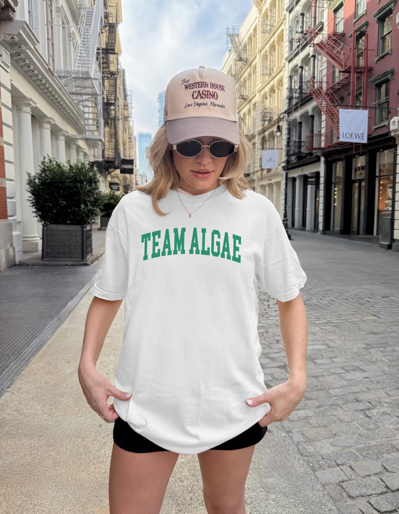 May include: A white t-shirt with the green text "TEAM ALGAE" printed in a collegiate font across the chest, paired with a beige and brown baseball cap featuring the text "The WESTERN HOUSE CASINO Las Vegas Nevada" and dark oval sunglasses.