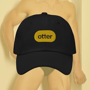 Puede incluir: Una gorra de béisbol negra con un parche ovalado amarillo que dice "otter" en letras negras.
