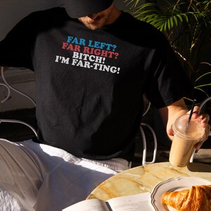 May include: Black t-shirt with the text "FAR LEFT? FAR RIGHT? BITCH! I'M FAR-TING!" in blue, red, and white. The shirt is worn by a person sitting at a table with a book, a croissant, and a drink.
