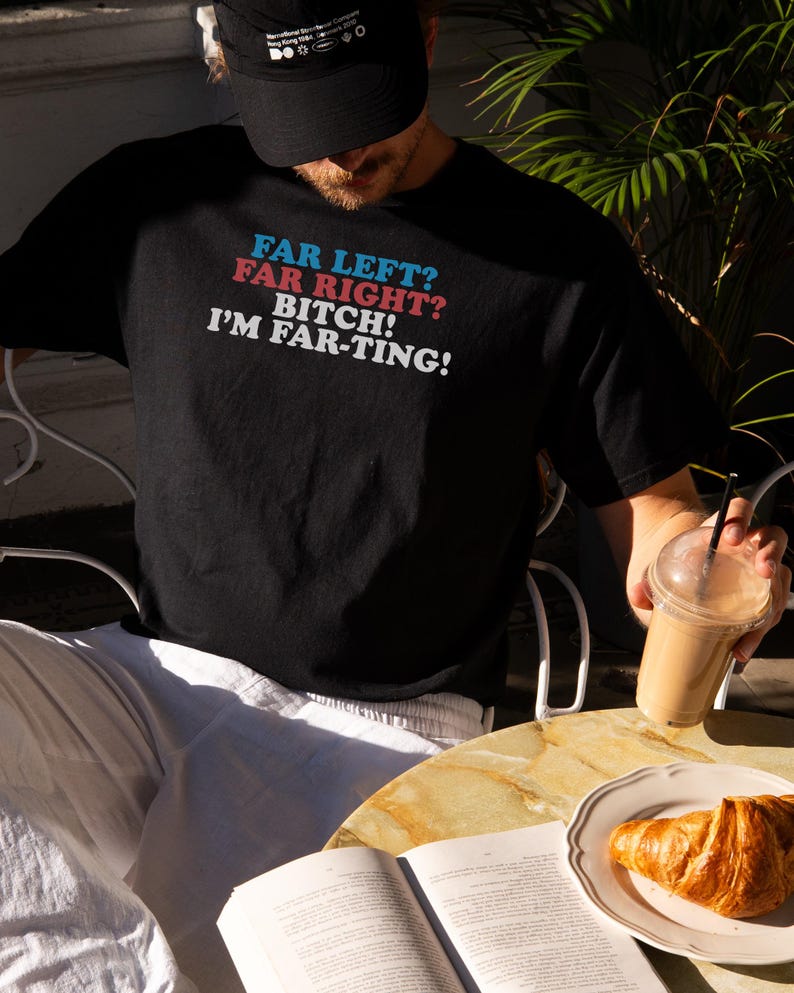 May include: Black t-shirt with the text "FAR LEFT? FAR RIGHT? BITCH! I'M FAR-TING!" in blue, red, and white. The shirt is worn by a person sitting at a table with a book, a croissant, and a drink.