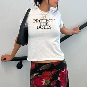 Puede incluir: Camiseta blanca con el texto negro "PROTECT THE DOLLS". La persona lleva un bolso negro y una falda con estampado floral con flores rojas sobre fondo negro. La persona tambi&eacute;n lleva gafas de sol.
