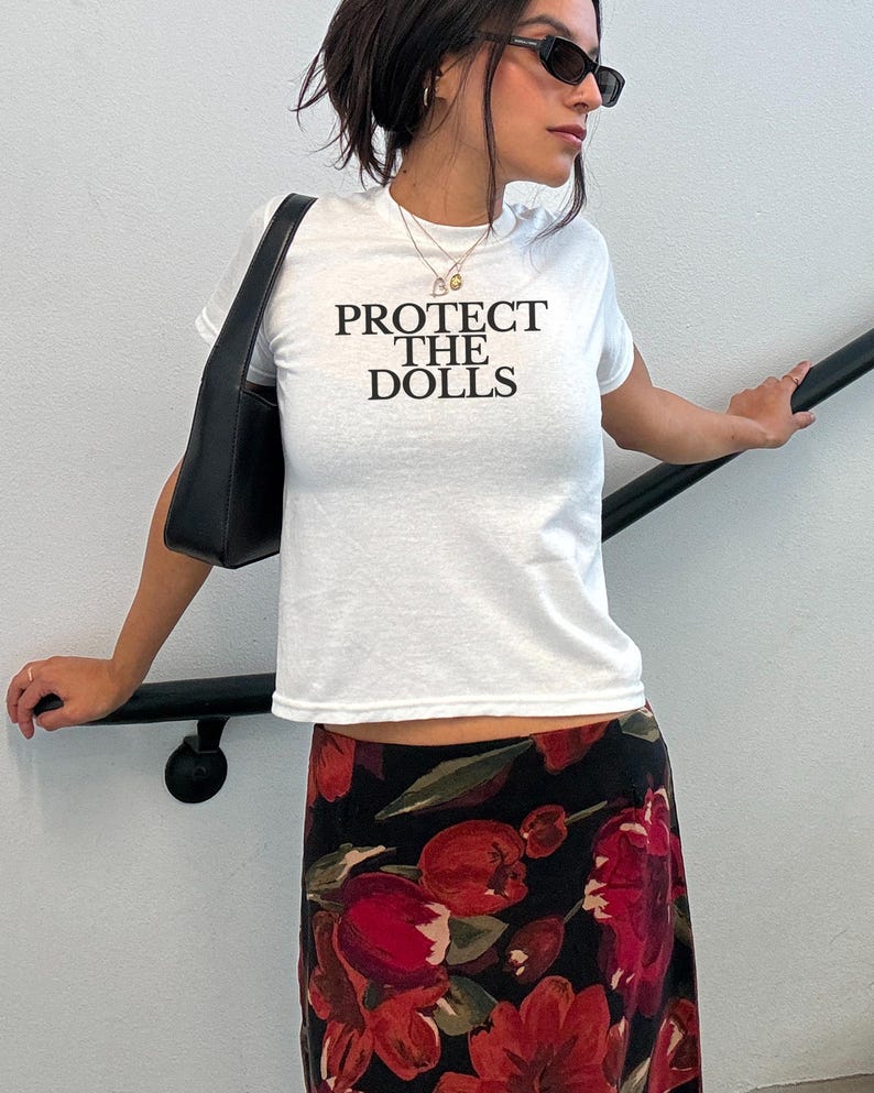 Puede incluir: Camiseta blanca con el texto negro "PROTECT THE DOLLS". La persona lleva un bolso negro y una falda con estampado floral con flores rojas sobre fondo negro. La persona tambi&eacute;n lleva gafas de sol.