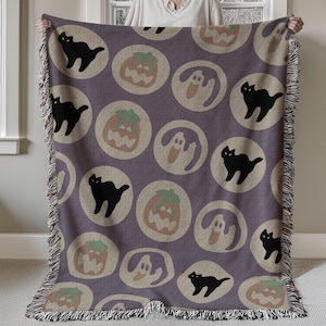 Op de afbeelding: Een paarse sprei met een herhaald patroon van zwarte katten, pompoenen en geesten in beige cirkels. De sprei heeft een gefranjerde rand en wordt door een persoon vastgehouden.
