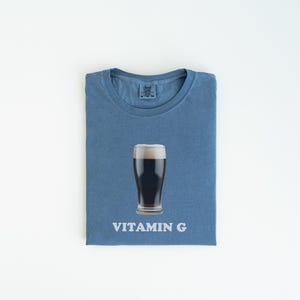 Pu&ograve; includere: Una maglietta blu con un bicchiere di birra scura e la scritta "VITAMIN G" stampata su di essa.