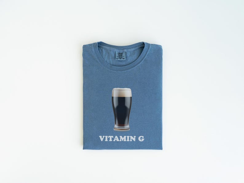 Pu&ograve; includere: Una maglietta blu con un bicchiere di birra scura e la scritta "VITAMIN G" stampata su di essa.