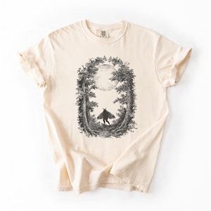 Puede incluir: Camiseta color crema con una ilustración en blanco y negro de una figura en un bosque con luna llena. El gráfico está enmarcado por árboles. La camiseta tiene mangas enrolladas y cuello redondo.