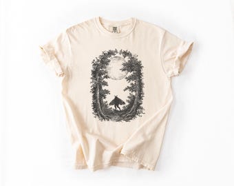 May include: Cream-colored t-shirt featuring a black and white illustration of a figure in a forest with a full moon. The graphic is framed by trees. The shirt has rolled-up sleeves and a crew neck.