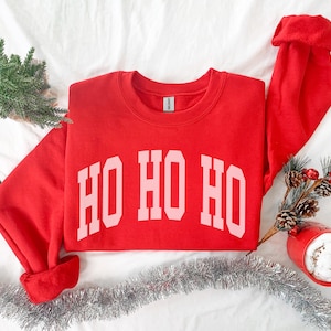 以下が含まれることがあります： 赤いクリスマスのトレーナーに、ピンクの文字で「HO HO HO」と書かれています。