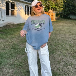 May include: A woman wearing a light blue t-shirt with the text "Zombie Island Pepper Plantation & Inn" and white pants. She is wearing a white baseball cap and sunglasses.