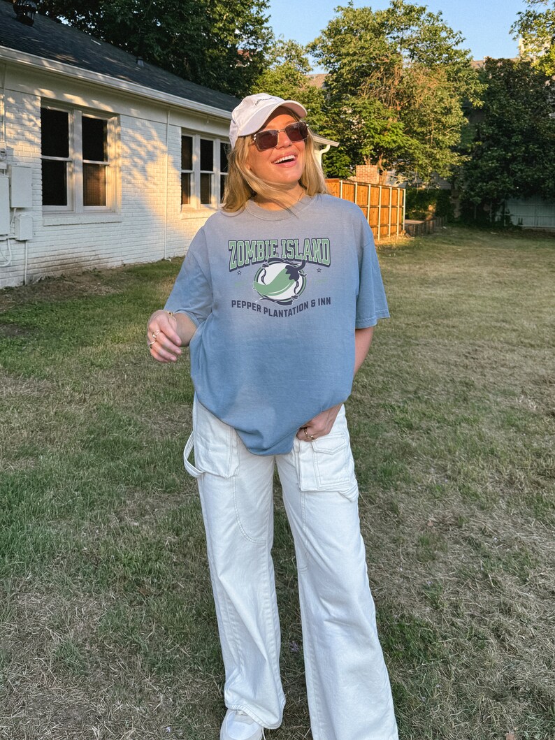 May include: A woman wearing a light blue t-shirt with the text "Zombie Island Pepper Plantation & Inn" and white pants. She is wearing a white baseball cap and sunglasses.