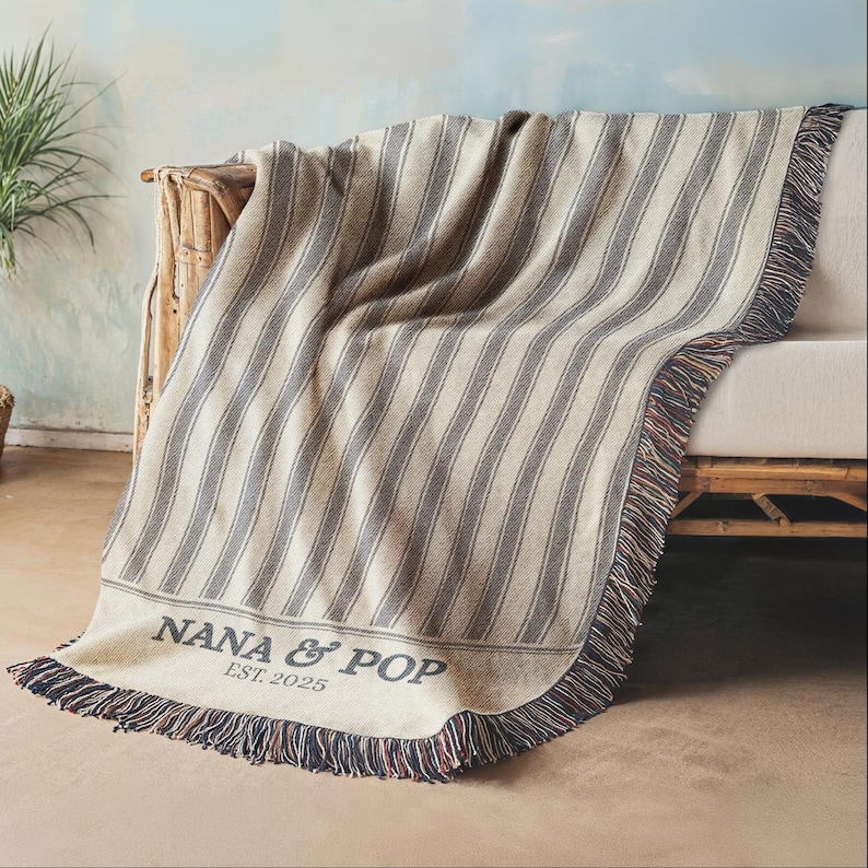 May include: A beige and gray striped throw blanket draped over a sofa. The blanket has a fringe border with red, white, and blue accents. The text "NANA & POP EST. 2025" is printed on the blanket.
