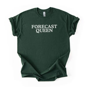 May include: Dark green t-shirt with the text "FORECAST QUEEN" in white block letters. The shirt has rolled-up sleeves and a crew neck. The fabric appears to be a soft, heathered material.