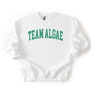 May include: A white crewneck sweatshirt featuring the text "TEAM ALGAE" printed in a bold, arched green font across the chest. The garment has long sleeves with ribbed cuffs and a ribbed hem, constructed from a solid white fabric material.