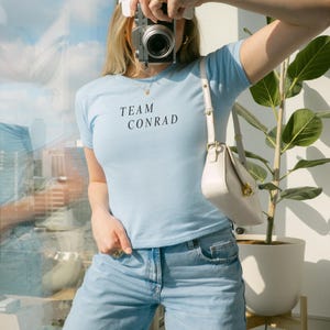 May include: A light blue t-shirt with the text "TEAM CONRAD" in black, paired with light blue jeans. A white purse and a white cap are also visible. The person is holding a silver camera.