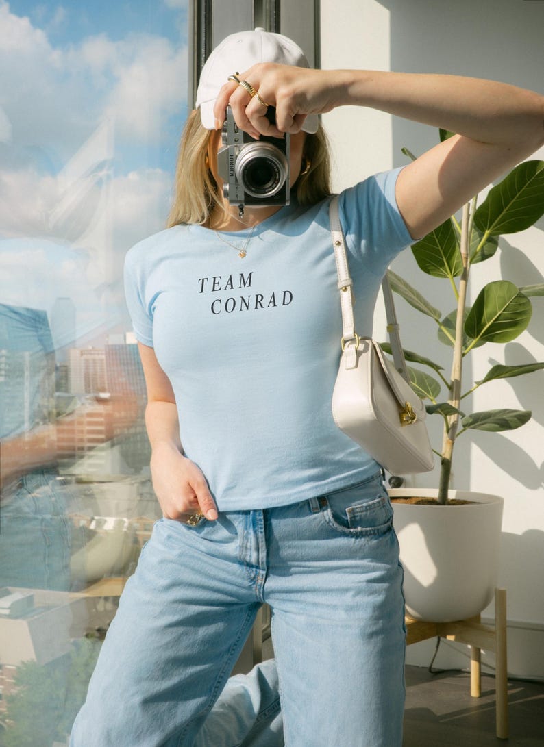 May include: A light blue t-shirt with the text "TEAM CONRAD" in black, paired with light blue jeans. A white purse and a white cap are also visible. The person is holding a silver camera.