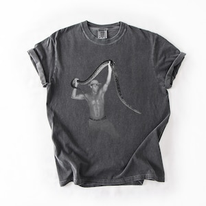 Puede incluir: Camiseta gris oscuro con una imagen en blanco y negro de una persona sosteniendo una serpiente. La imagen está centrada en la camiseta, con los brazos de la persona levantados, sosteniendo la serpiente. La camiseta tiene cuello redondo y mangas cortas.