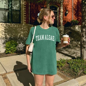 May include: A forest green short-sleeve crewneck t-shirt featuring the text "TEAM ALGAE" printed in white across the chest in a collegiate-style font. The shirt has a relaxed fit and is made of a solid-colored cotton fabric with a smooth texture.