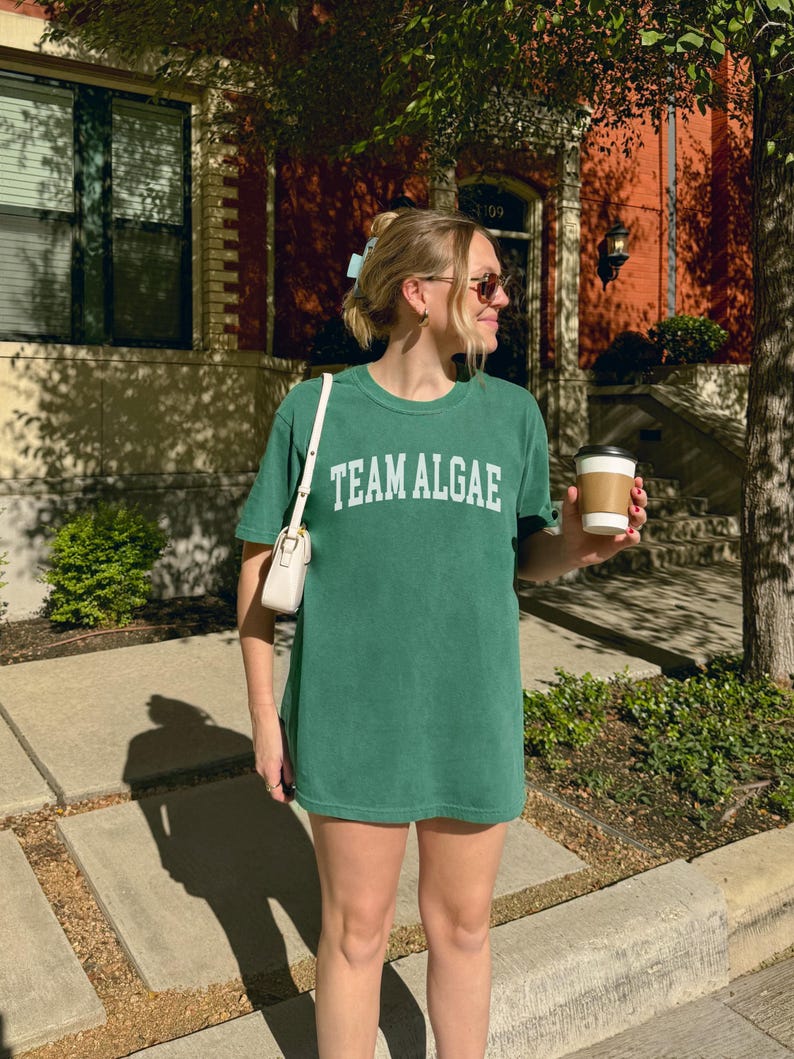 May include: A forest green short-sleeve crewneck t-shirt featuring the text "TEAM ALGAE" printed in white across the chest in a collegiate-style font. The shirt has a relaxed fit and is made of a solid-colored cotton fabric with a smooth texture.