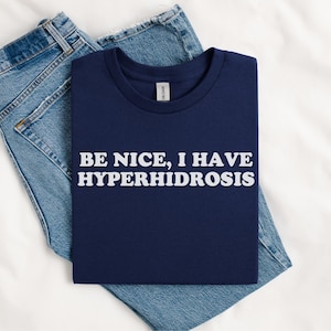 以下が含まれることがあります： 白い文字で「BE NICE, I HAVE HYPERHIDROSIS」と書かれた紺色のTシャツ。Tシャツは折り畳まれて、青いジーンズの上に置かれています。