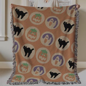 May include: A Halloween-themed throw blanket with a repeating pattern of black cats, pumpkins, and ghosts in white circles on a tan backdrop. The blanket has a fringed edge.