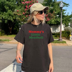 May include: Black t-shirt featuring the text "Mommy's Little Meatball" in red and green. The casual tee is suitable for everyday wear. The image is taken outdoors on a sunny day.