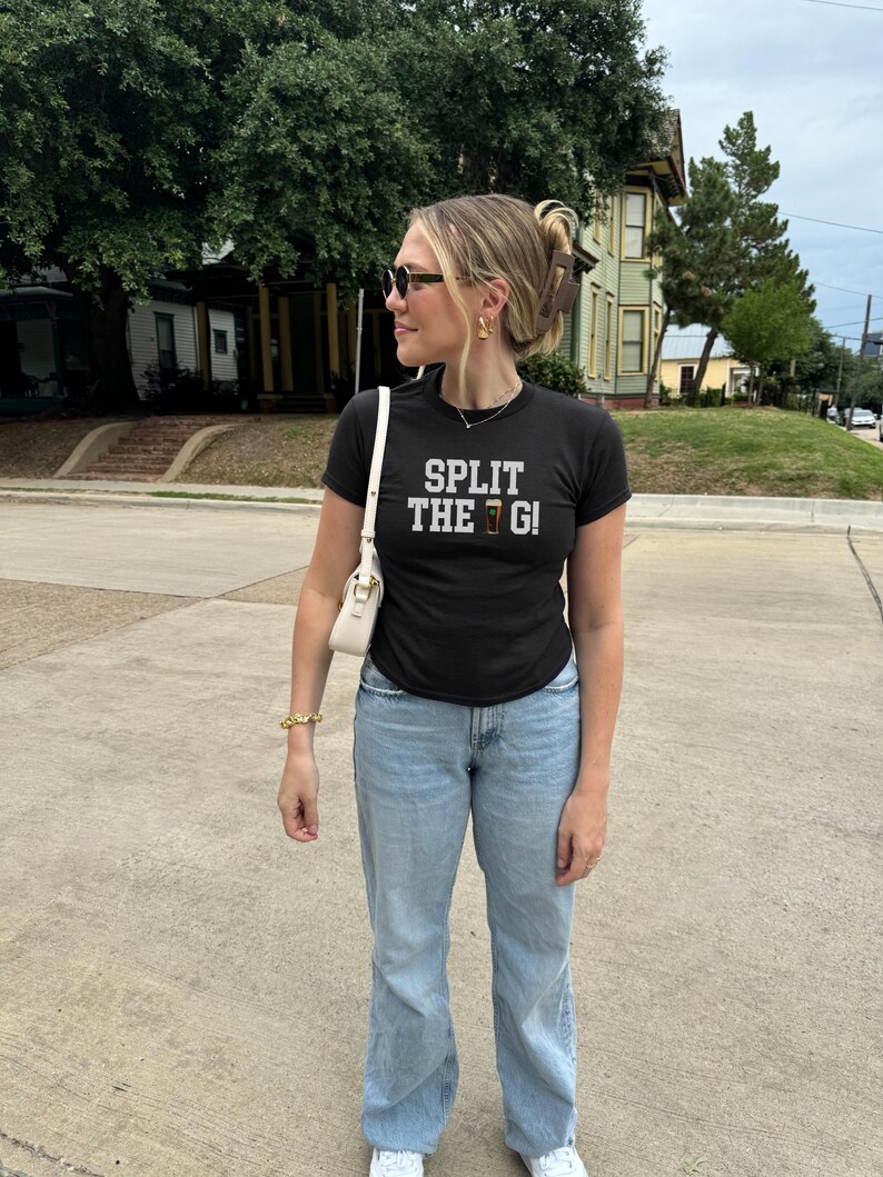 Pu&ograve; includere: T-shirt nero con la scritta "SPLIT THE G!" in bianco e marrone. La maglietta &egrave; abbinata a jeans azzurri e scarpe da ginnastica bianche. Una borsa bianca e gioielli dorati completano il look. La persona indossa occhiali da sole.
