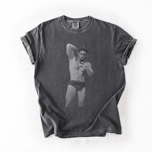 May include: A charcoal gray short-sleeve t-shirt featuring a centered black and white graphic print of a person wearing glasses and underwear while taking a mirror selfie. The shirt has a crew neckline and a washed, vintage-style fabric texture.