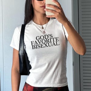 Op de afbeelding: Wit T-shirt met de tekst "God's Favorite Bisexual" gedrukt op de voorkant.
