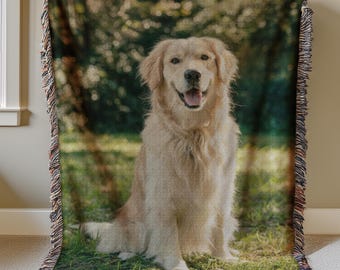 May include: A woven throw blanket featuring a golden retriever dog sitting in a grassy field. The dog has a happy expression. The background shows a blurred green forest. The blanket has a fringed edge.