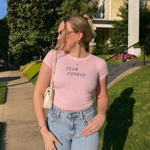 May include: A light pink, short-sleeved t-shirt with the text "TEAM CONRAD" in black. The shirt is paired with light blue jeans and a white purse. The person is wearing sunglasses and jewelry.