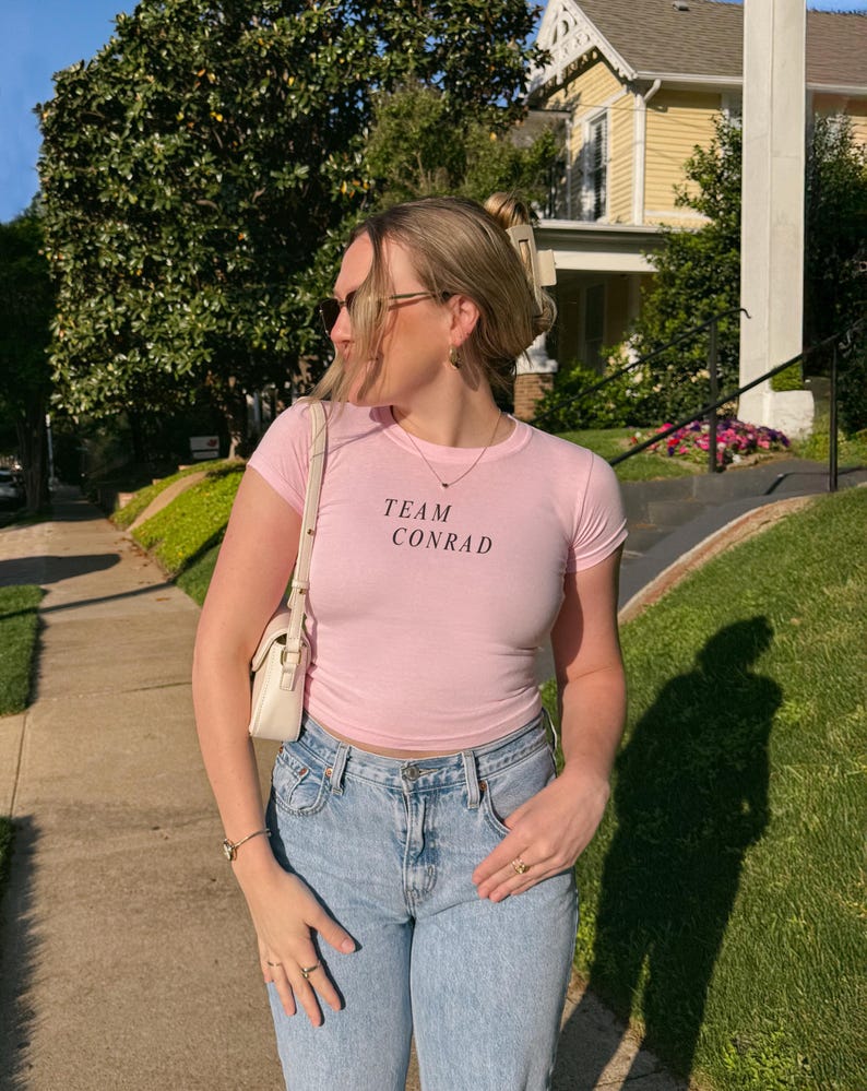 May include: A light pink, short-sleeved t-shirt with the text "TEAM CONRAD" in black. The shirt is paired with light blue jeans and a white purse. The person is wearing sunglasses and jewelry.
