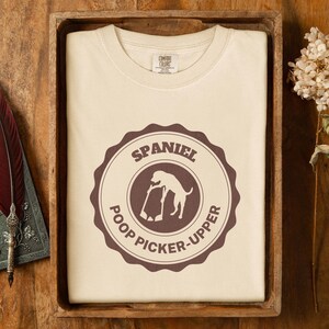 May include: A white t-shirt with a brown circle design. The circle has the text "Spaniel Poop Picker-Upper" and a silhouette of a dog picking up poop.