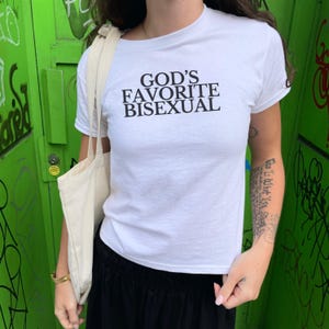 Op de afbeelding: Wit T-shirt met zwarte tekst die "God's Favorite Bisexual" zegt.