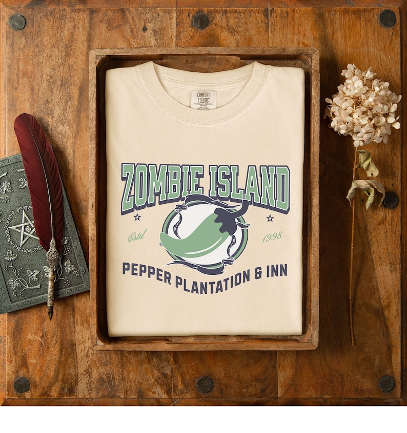 May include: A beige t-shirt with a green chili pepper graphic and the text "Zombie Island Pepper Plantation & Inn" in navy blue and white. The text "Estd 1998" is also included.