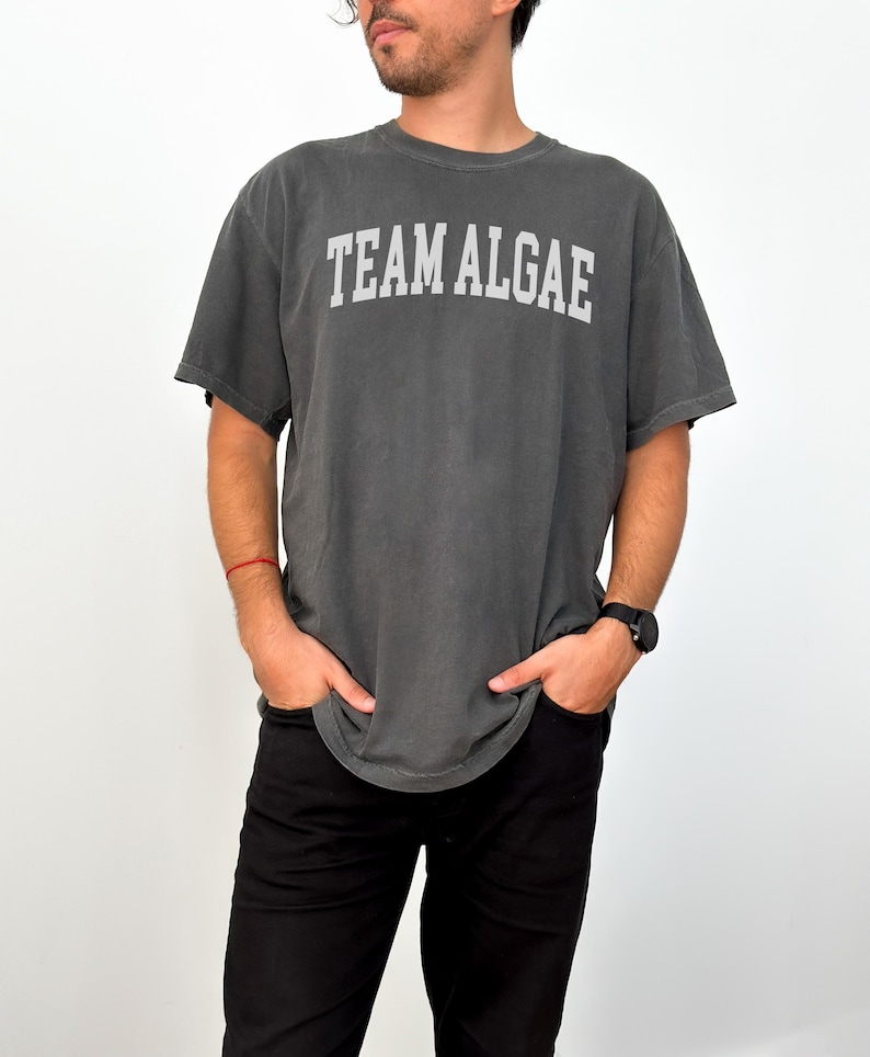May include: A charcoal grey short-sleeve t-shirt featuring the text "TEAM ALGAE" printed in a light grey, arched collegiate-style font across the chest. The garment has a crew neckline and a relaxed fit, paired with solid black trousers.