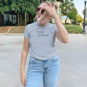 May include: A light gray t-shirt with the text "TEAM CONRAD" in black. The person is wearing light blue jeans and sunglasses. A gold bracelet is visible on the left wrist.